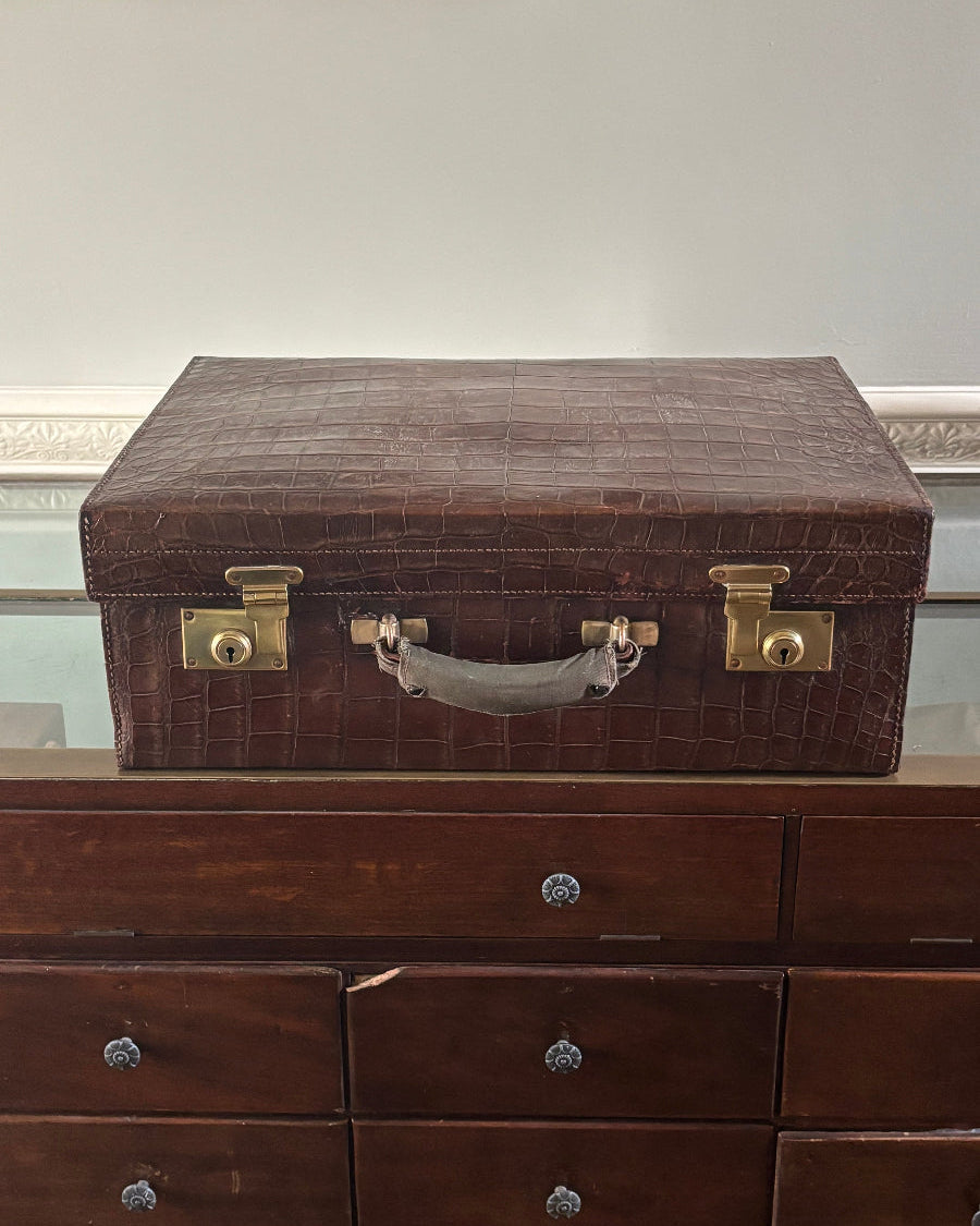 A dark brown embossed crocodile-patterned briefcase with brass hardware and a gray handle, shown standing vertically next to a dark wood drawer cabinet.