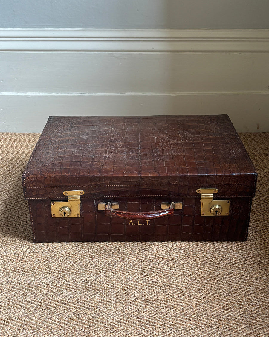 A dark brown embossed leather trunk or briefcase with brass latches and a handle, sitting on a woven beige carpet against a white baseboard and gray wall.