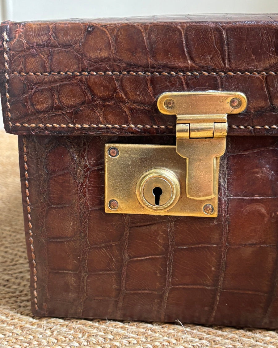 Close-up of a brown crocodile-embossed leather case featuring a brass clasp and keyhole lock, set against a neutral woven background.
