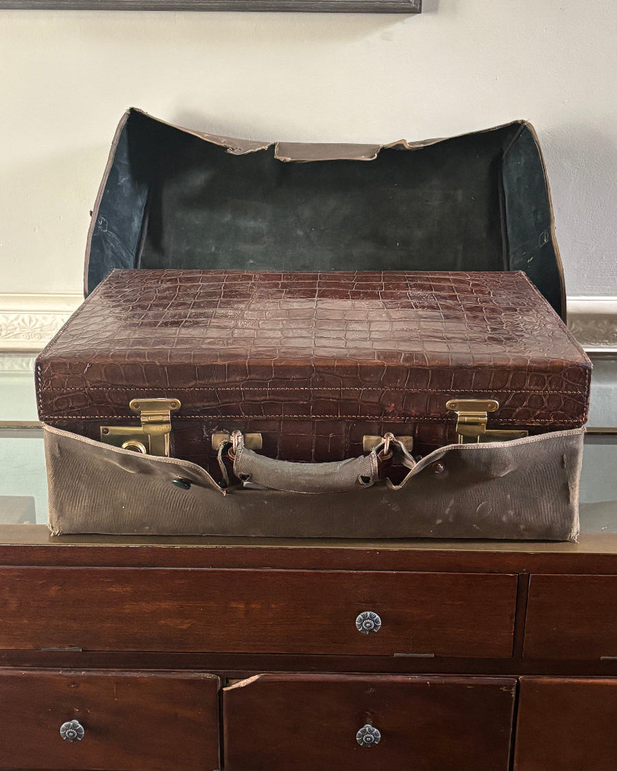 A vintage brown crocodile-embossed briefcase or suitcase with brass hardware, shown open and standing vertically next to a dark wooden drawer cabinet.