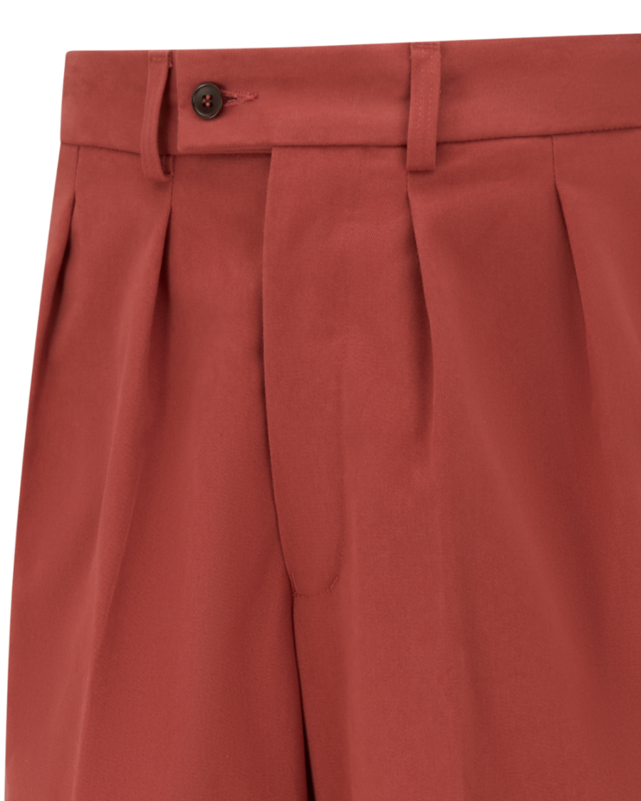 Close-up of pleated trousers in a muted terracotta or rust red color, showing the waistband, button, belt loops, and front pleats against a plain white background.