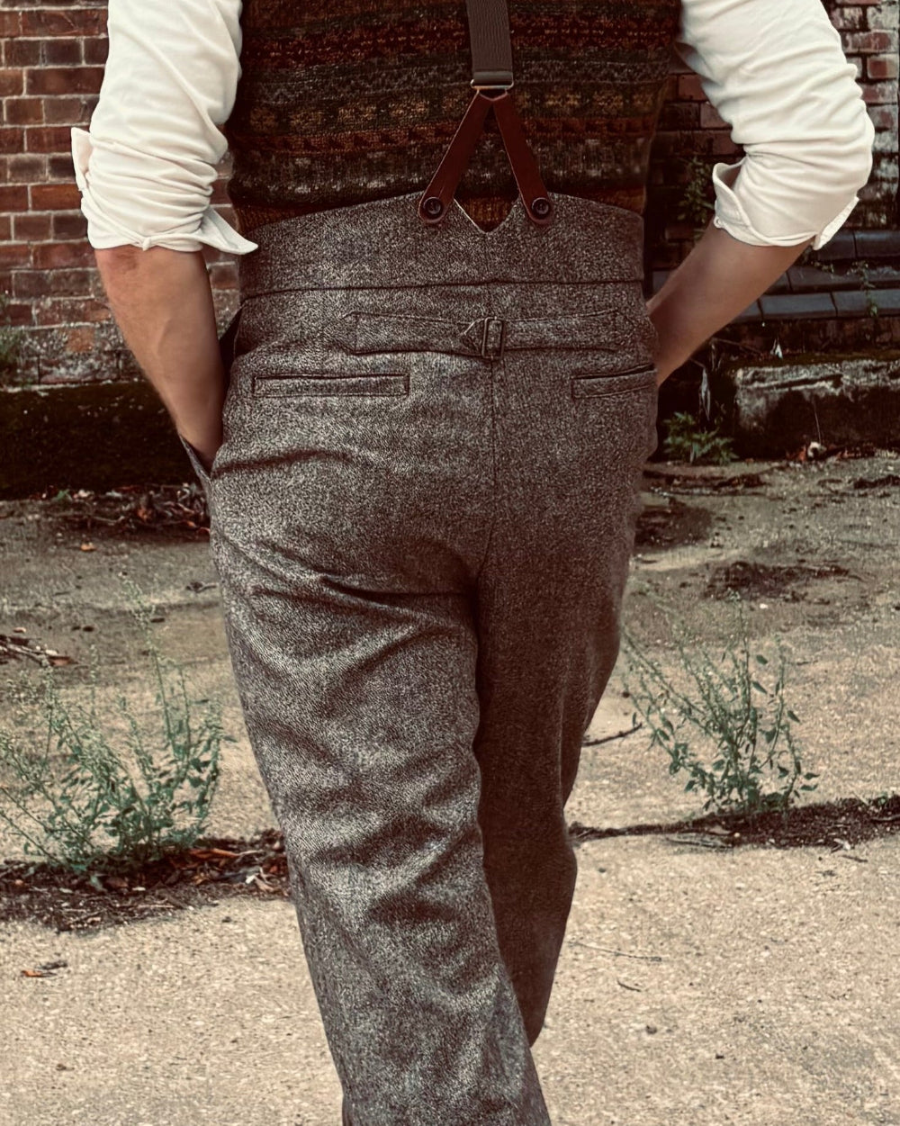 A man viewed from behind wearing a brown tweed waistcoat with brown suspenders, a cream shirt, gray herringbone trousers, and a brown flat cap, standing in front of a red brick wall.
