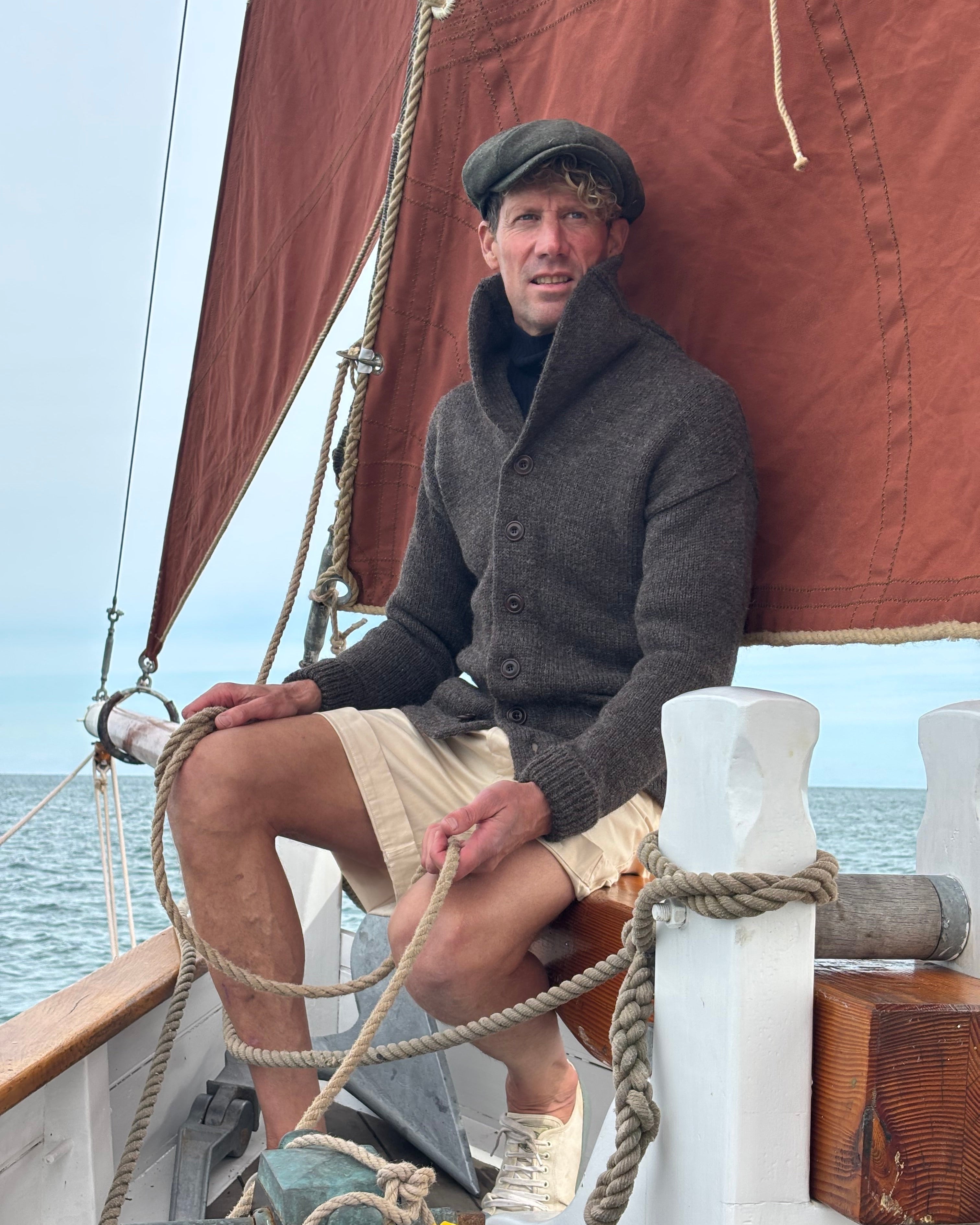 A man wearing a dark gray button-up sweater, beige shorts, and a dark flat cap sits on a wooden boat deck, holding a thick rope near the rigging under a brown canvas sail.