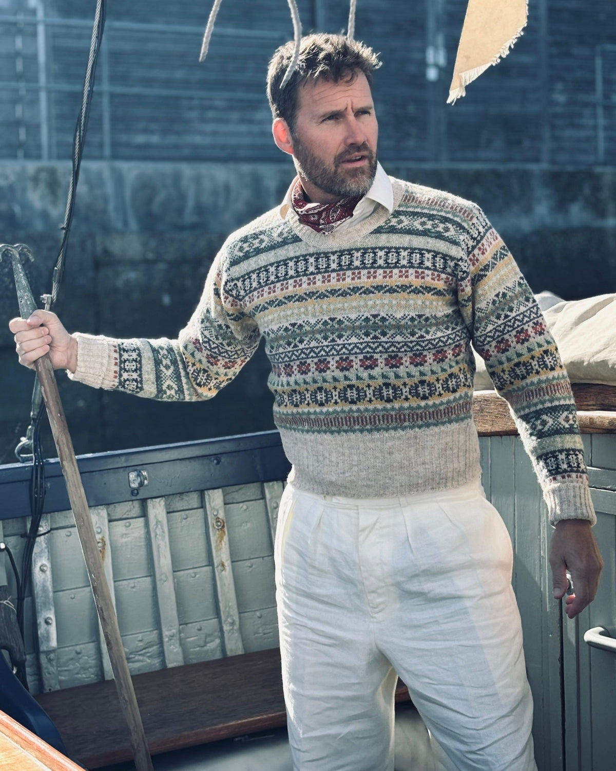 A man wearing a cream and navy Fair Isle patterned knit sweater, white trousers, and a red neck scarf, standing on a boat deck.