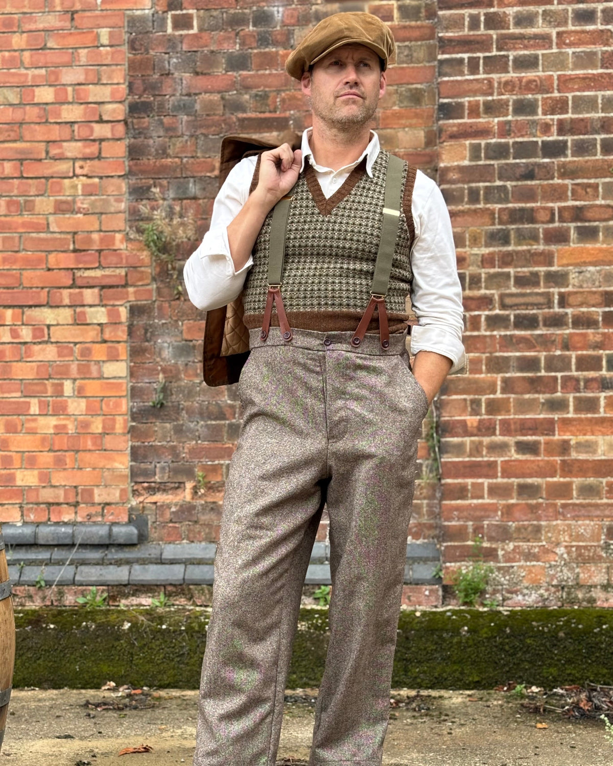 A man wearing a vintage-style tweed outfit stands in front of a brick wall. He is wearing brown tweed trousers, a brown tweed vest over a white shirt, a brown flat cap, and brown leather shoes, with a brown leather bag slung over his shoulder.