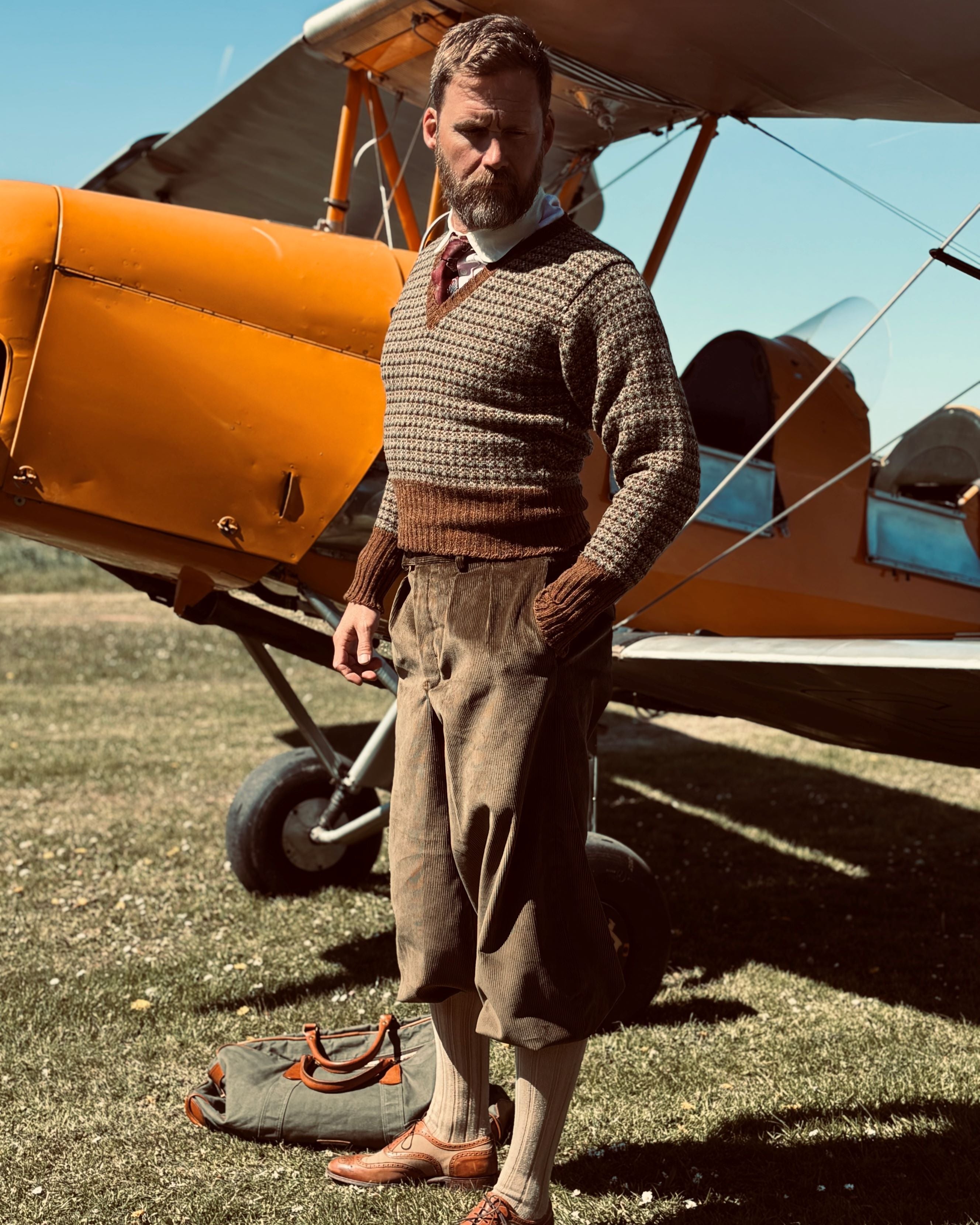 Man in vintage clothing standing next to a biplane on a grassy field.