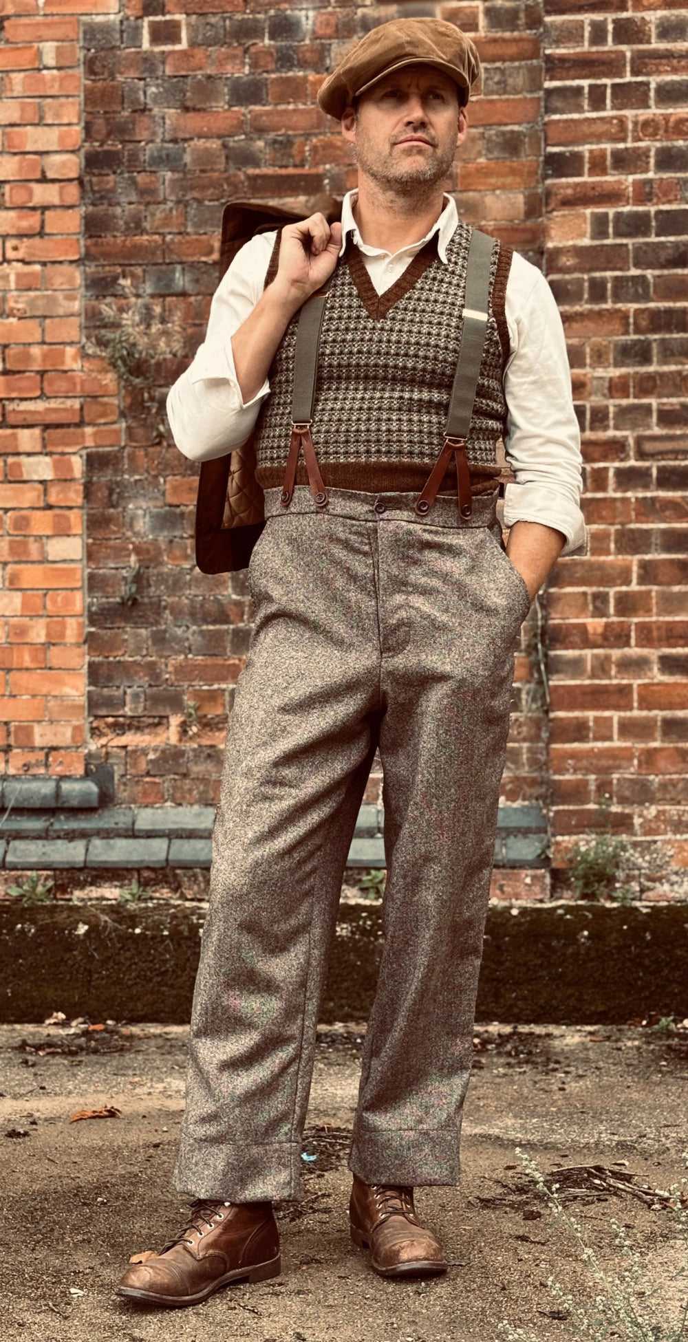 A man wearing a vintage-style tweed outfit with patterned overalls, a houndstooth vest, a cream shirt, a brown flat cap, and brown leather shoes, standing in front of a brick wall.