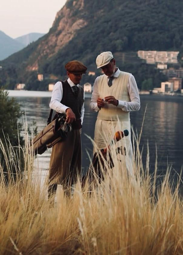 Two men in vintage-style clothing standing in tall golden grass by a lake, with mountains and buildings in the background. Playing golf with Hickory golf clubs.