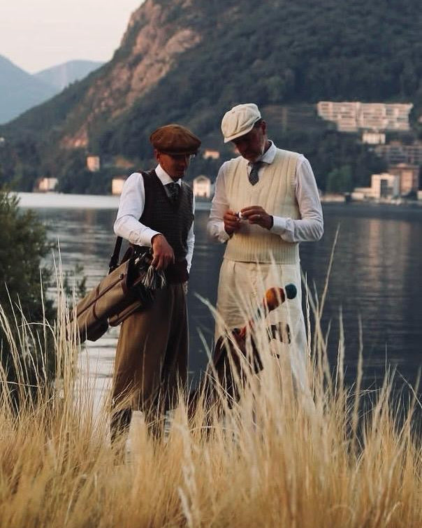 Two men in vintage-style clothing standing in tall golden grass by a lake, with mountains and buildings in the background. Playing golf with Hickory golf clubs.