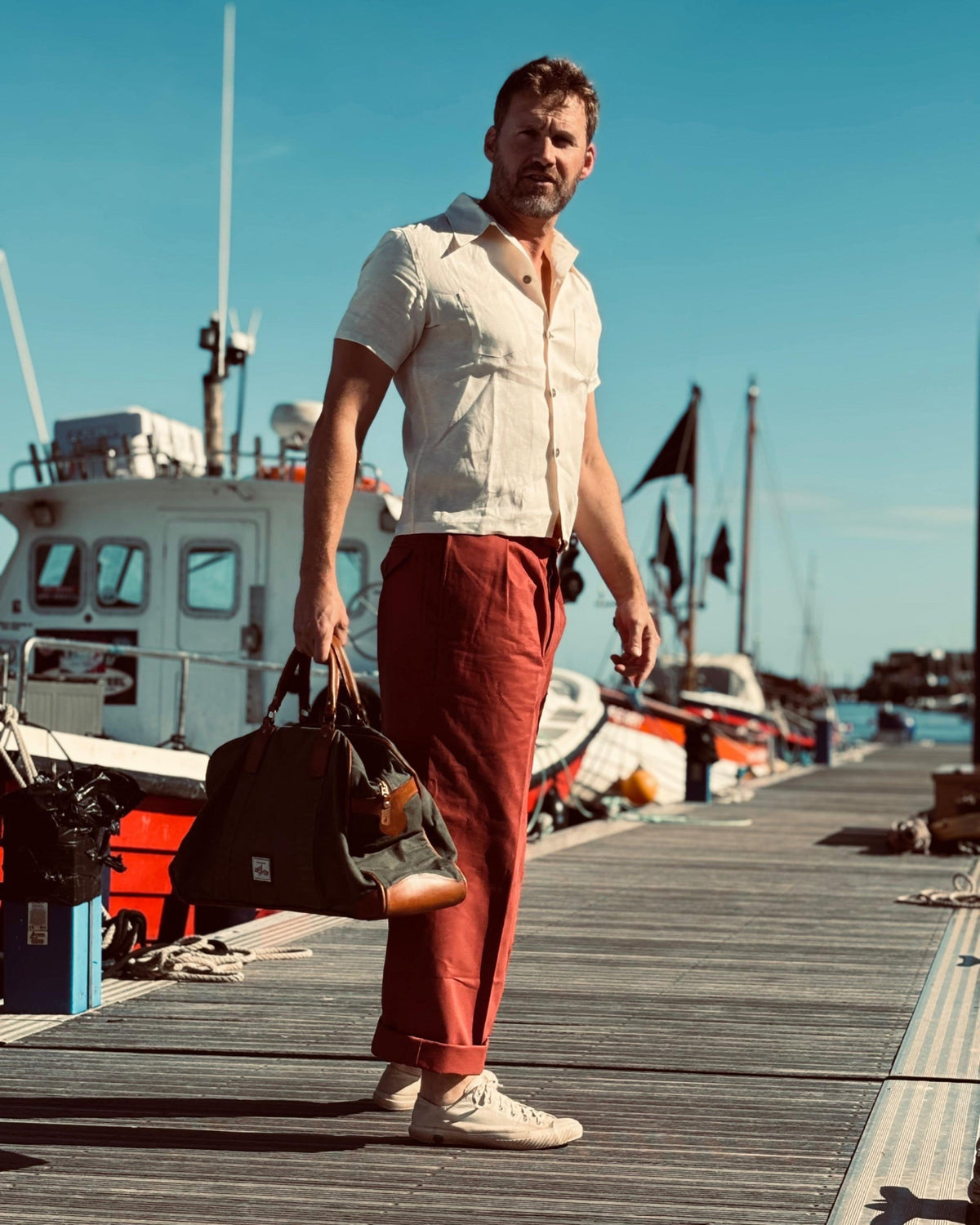 A man walks along a wooden dock holding a black duffel bag, wearing a white short-sleeve button-up shirt, 1930s rust-red trousers, and white trainers, with boats and a blue sky in the background.