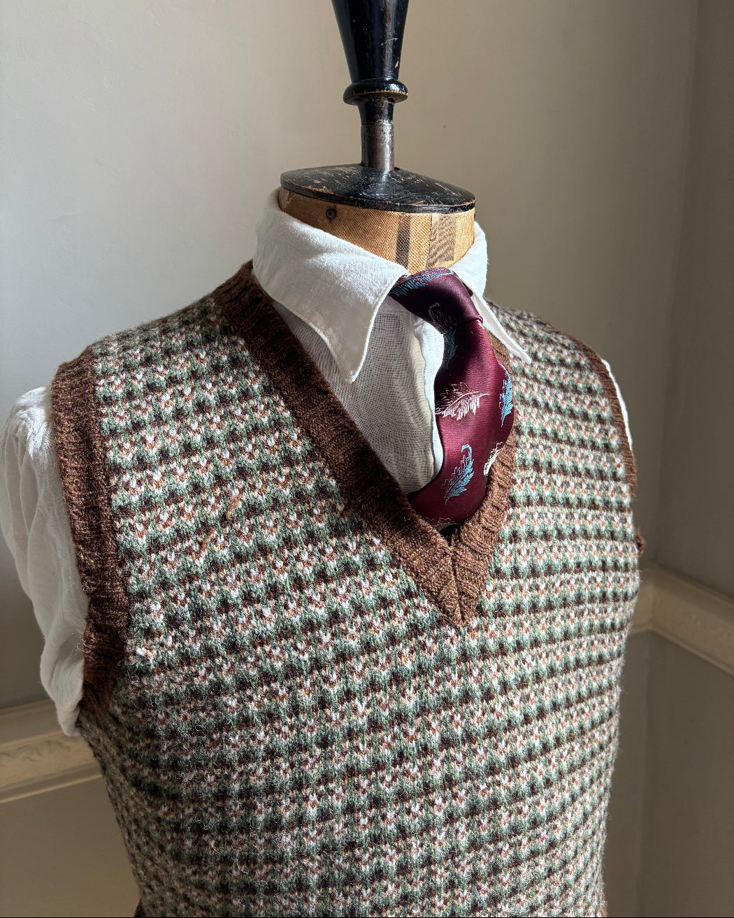 A knit sweater vest with a brown and beige houndstooth pattern, styled on a dress form with a white collared shirt and a burgundy paisley necktie.