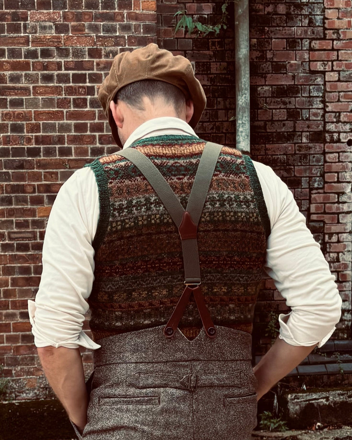 A man viewed from behind wearing a brown tweed waistcoat with brown suspenders, a cream shirt, gray herringbone trousers, and a brown flat cap, standing in front of a red brick wall.