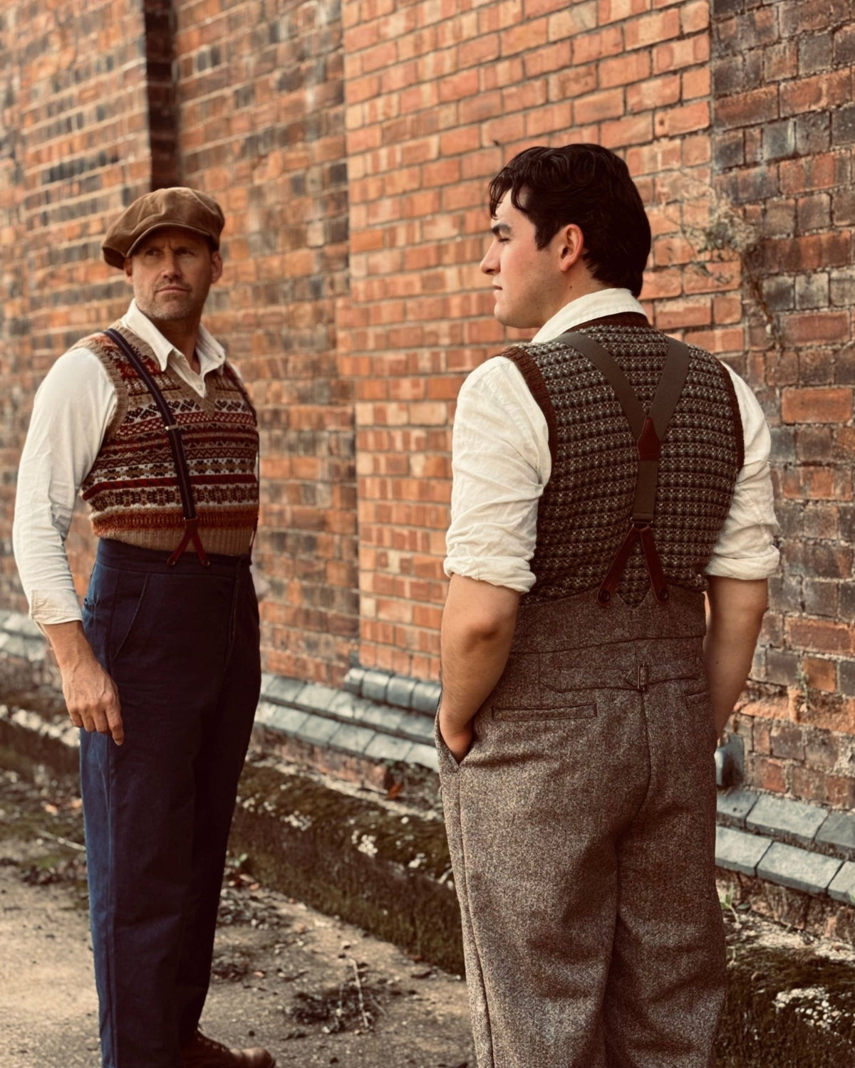 Two men in vintage-style tweed and knit clothing standing in front of a red brick wall, one facing forward wearing a patterned sweater vest and flat cap, and the other shown from the back wearing a textured vest and gray tweed overalls.
