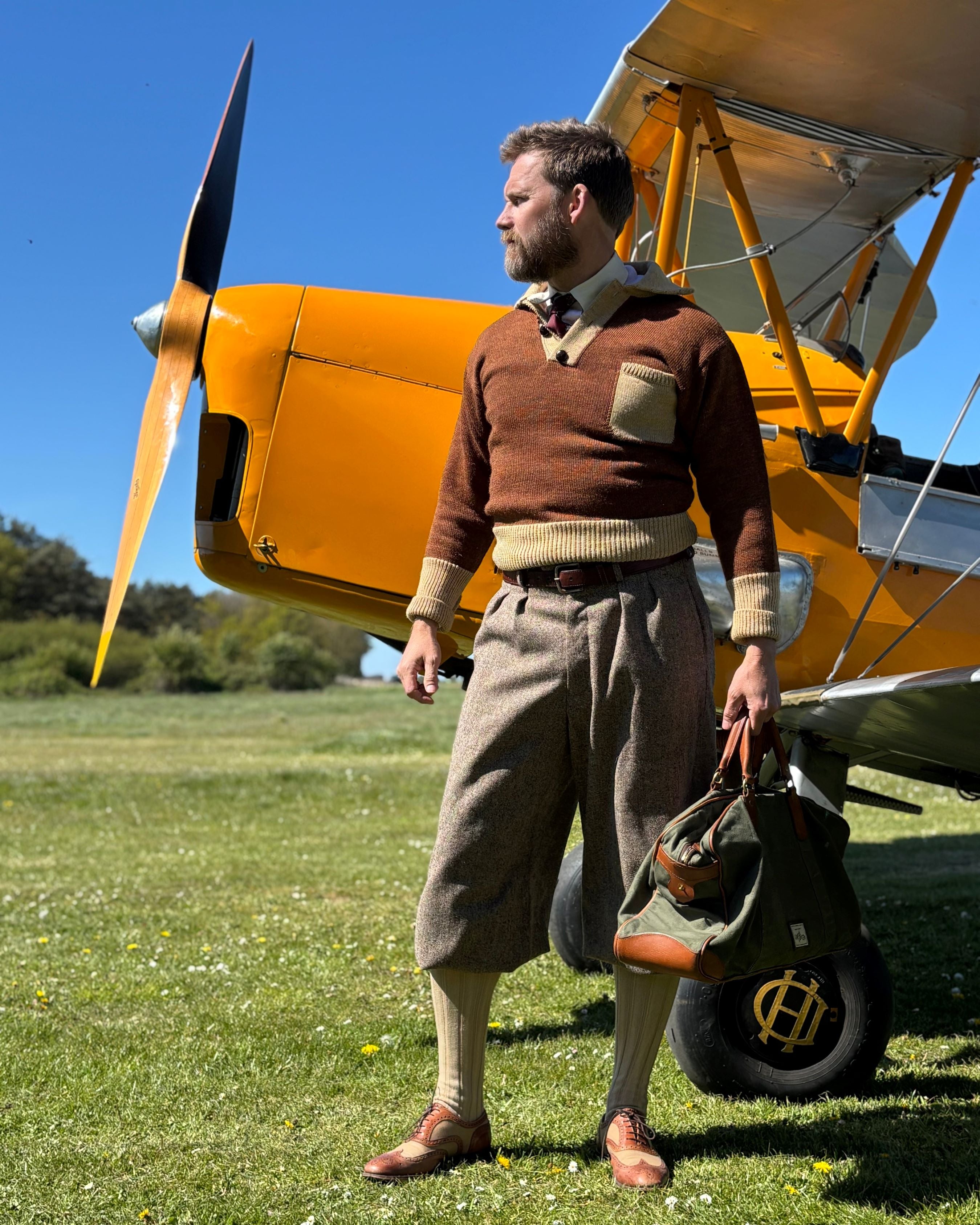 Man in vintage attire standing next to a yellow biplane on a grassy field.