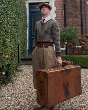 Man in vintage attire holding a brown suitcase outside a brick building.