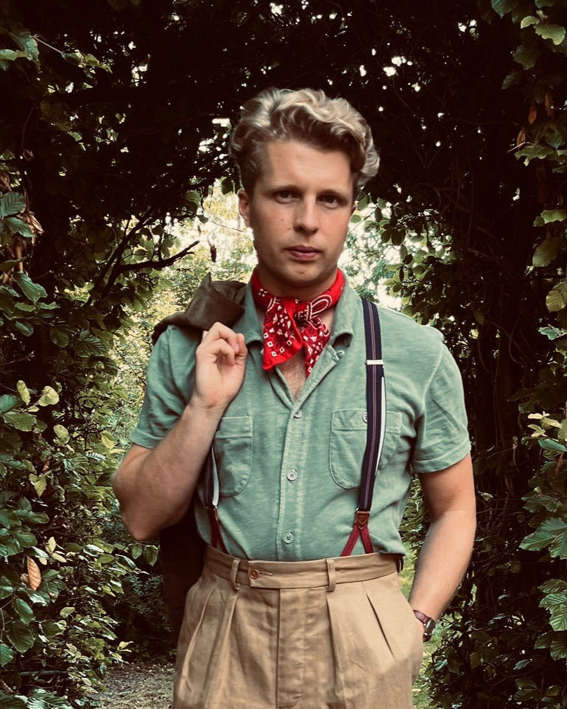 Man in a British made green shirt with suspenders and a red bandana standing in a forest.