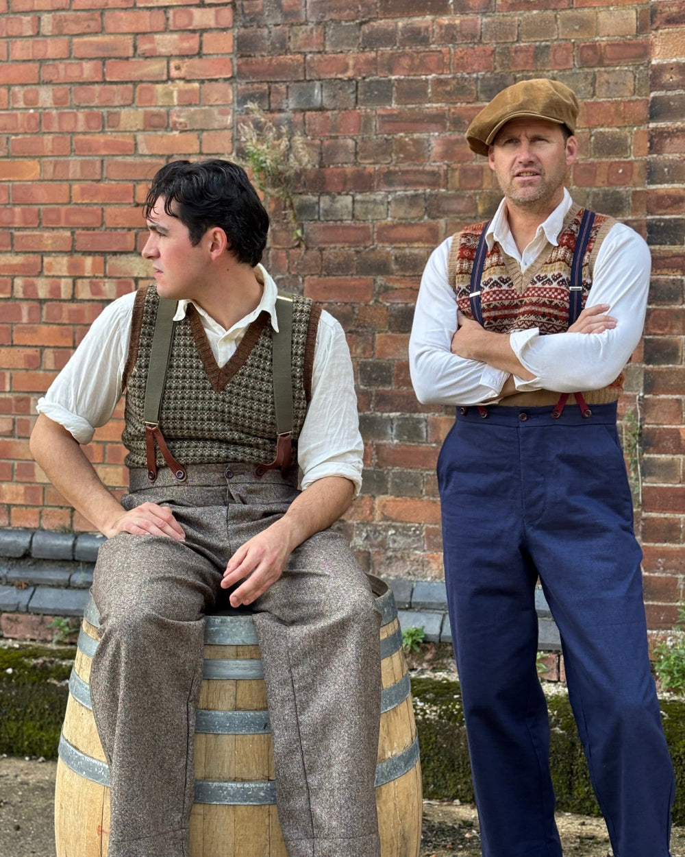 Two men dressed in vintage workwear standing and sitting in front of a brick wall. One man sits on a wooden barrel wearing a white shirt, brown tweed waistcoat and trousers.