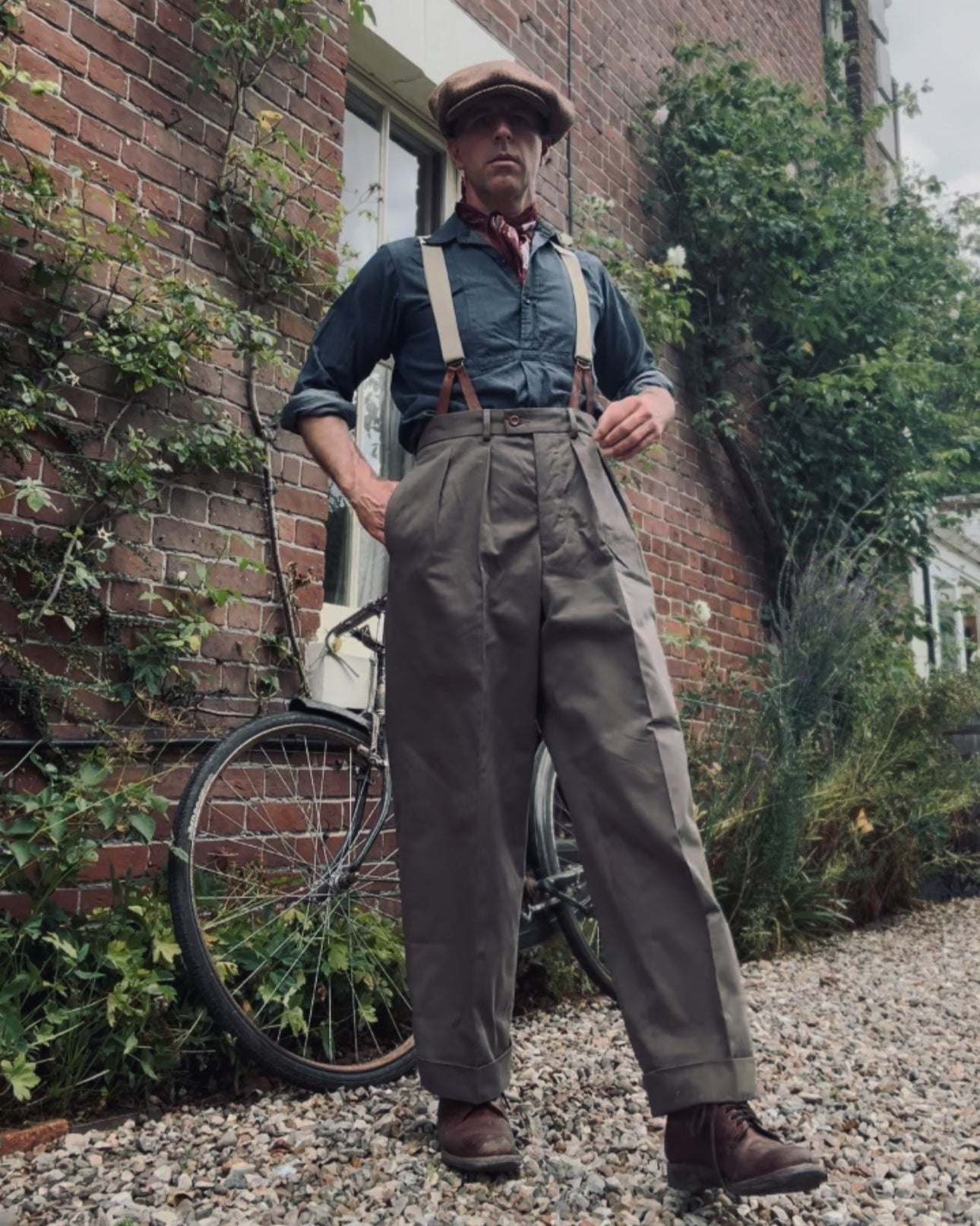 Man in suspenders standing next to a bicycle against a brick wall with plants.