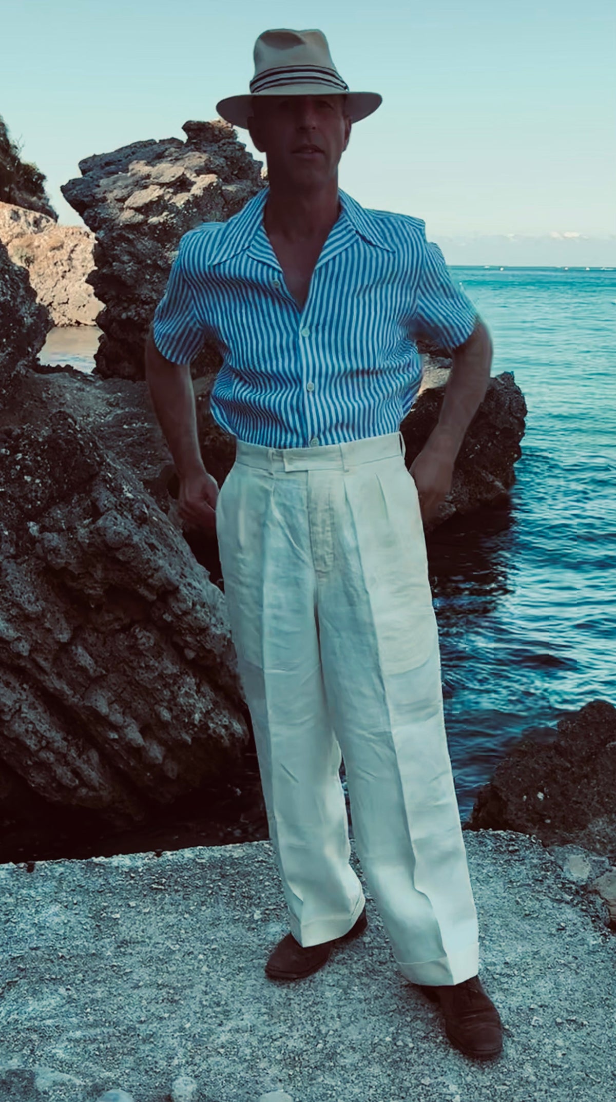 A man standing outdoors by the water wearing a light blue and white striped short-sleeve button-up shirt, white wide-leg trousers, a beige fedora hat, and dark brown shoes.
