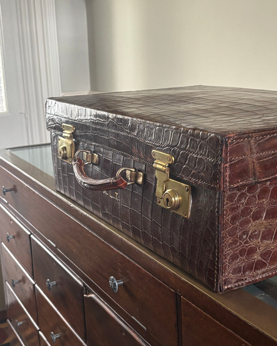 A dark brown crocodile-embossed leather briefcase or trunk with gold-tone hardware, shown resting on top of a dark wood dresser in an indoor room setting.