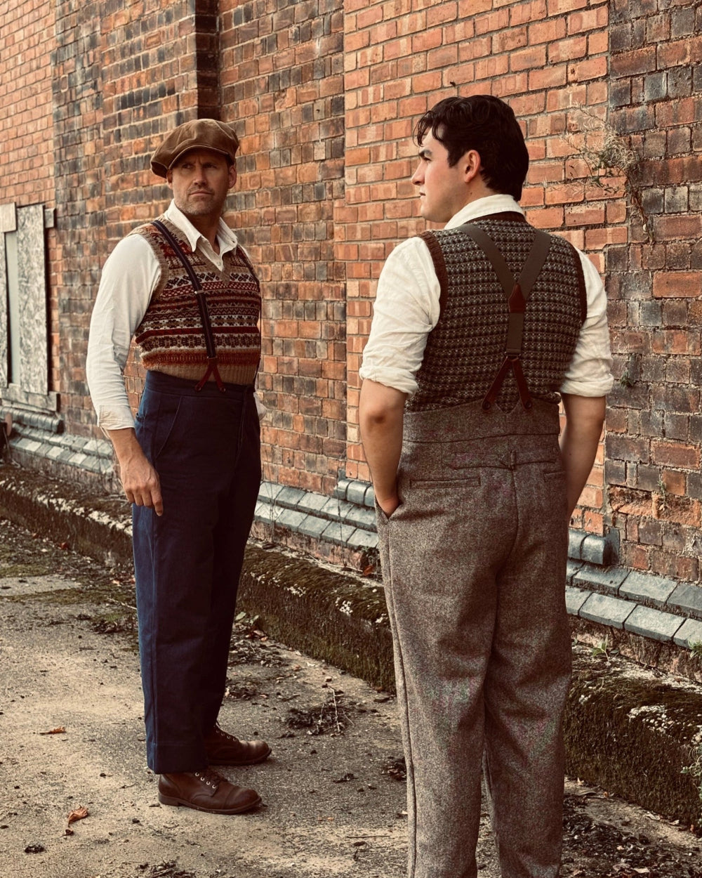 Two men standing outdoors against a red brick wall, wearing vintage-style tweed overalls and patterned knit vests. One man faces forward wearing navy trousers and a brown hat, while the other faces away showing the back of his gray tweed overalls.