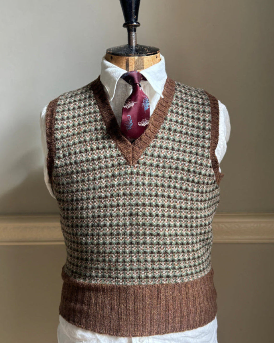 A brown and beige houndstooth knit sweater vest displayed on a mannequin torso, styled with a white collared shirt and a burgundy paisley necktie against a neutral beige wall.