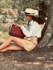 Woman sitting under a tree holding a book, wearing a hat and a scarf in vintage clothing.