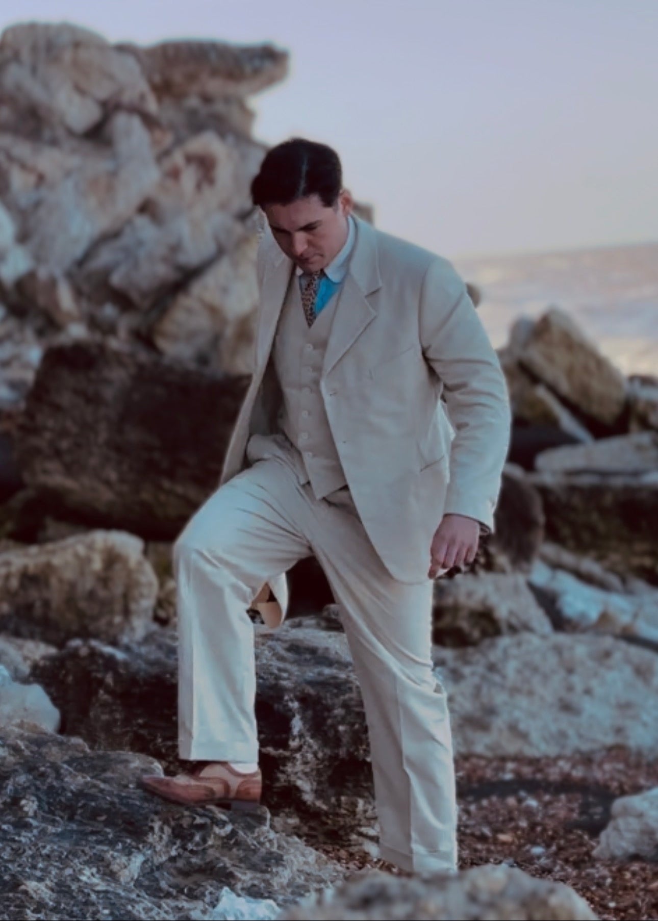 Man in a beige suit standing on rocky terrain with a scenic background