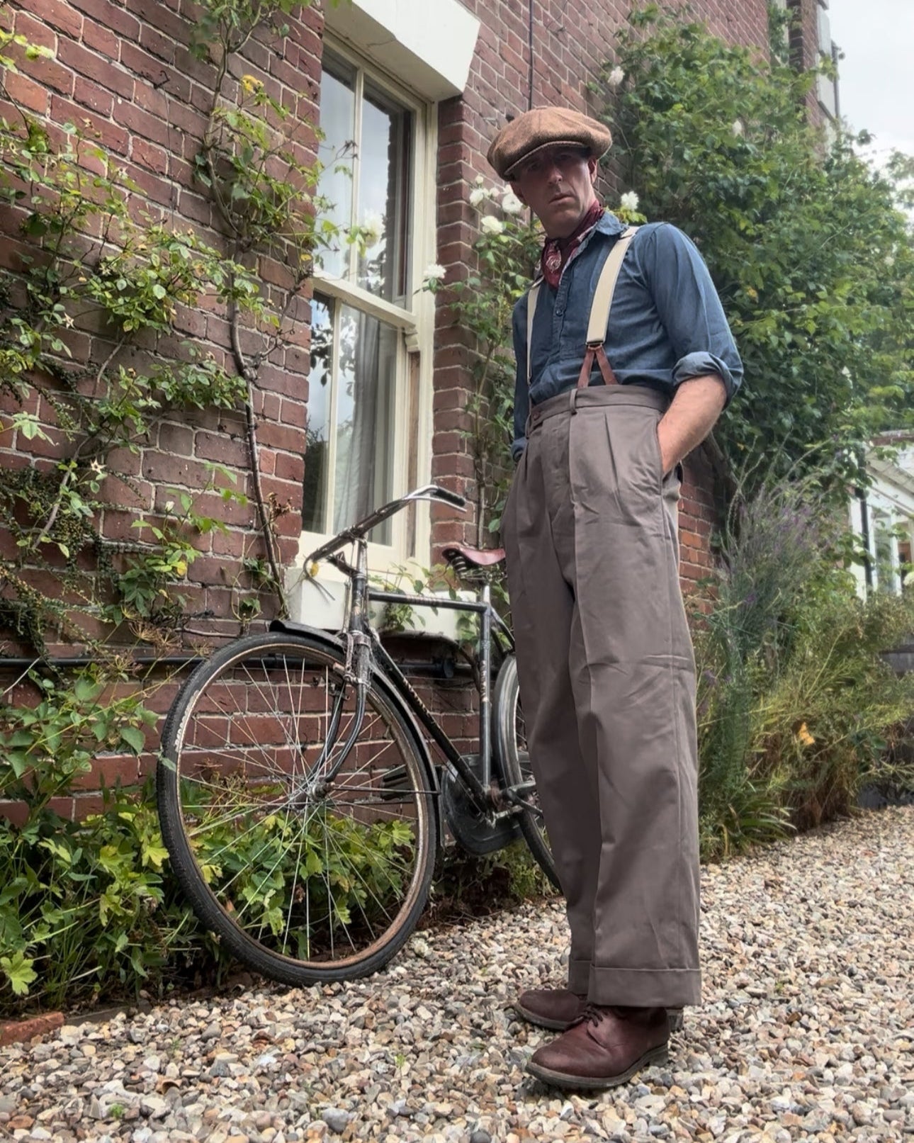 Man in vintage attire standing next to a bicycle against a brick wall with greenery.