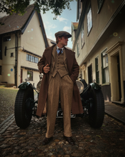 Man in vintage attire standing next to a classic car on a street.