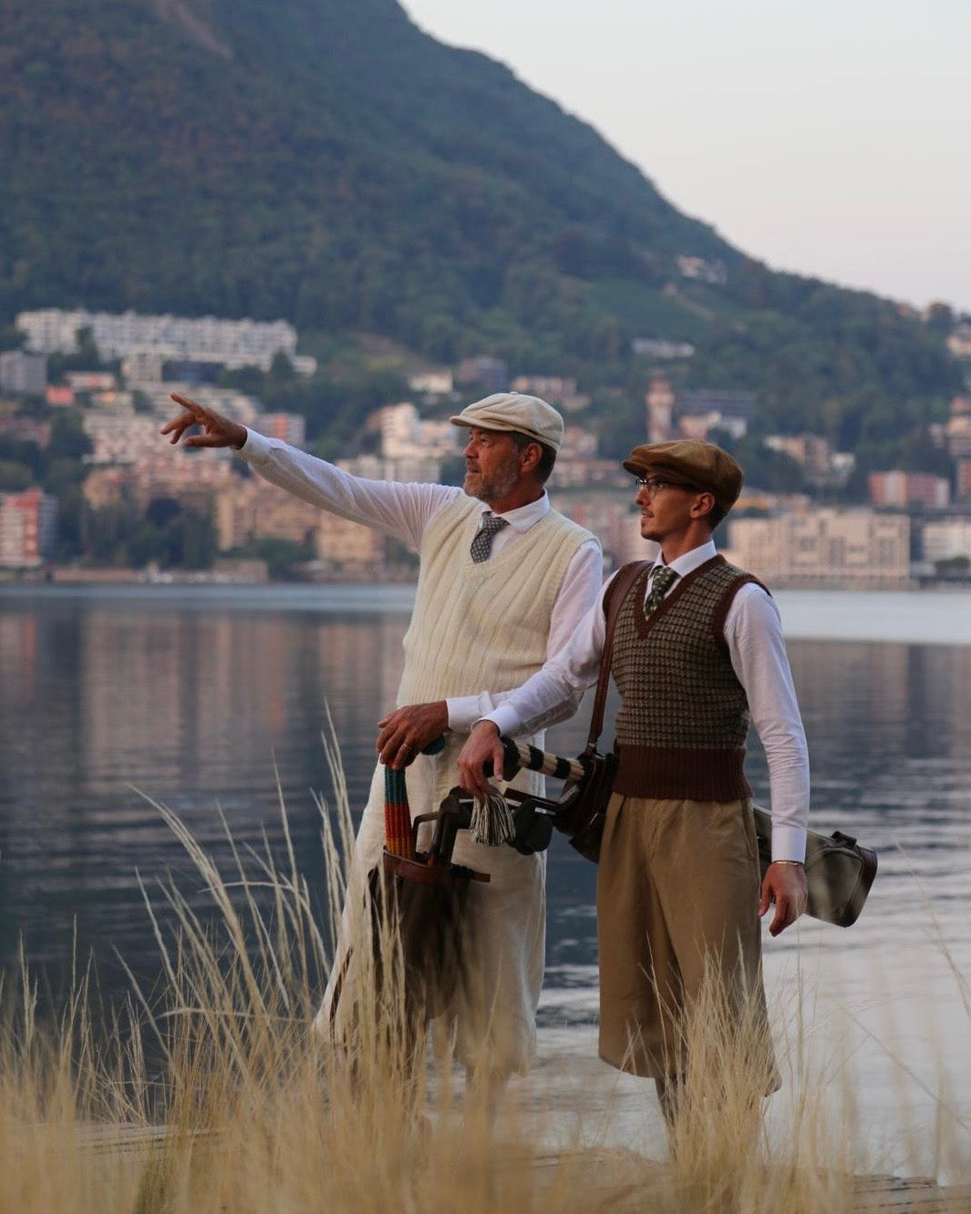 Two men dressed in vintage golfing attire standing by a lake with a mountain and town in the background, one pointing toward the landscape while holding hickory golf clubs.