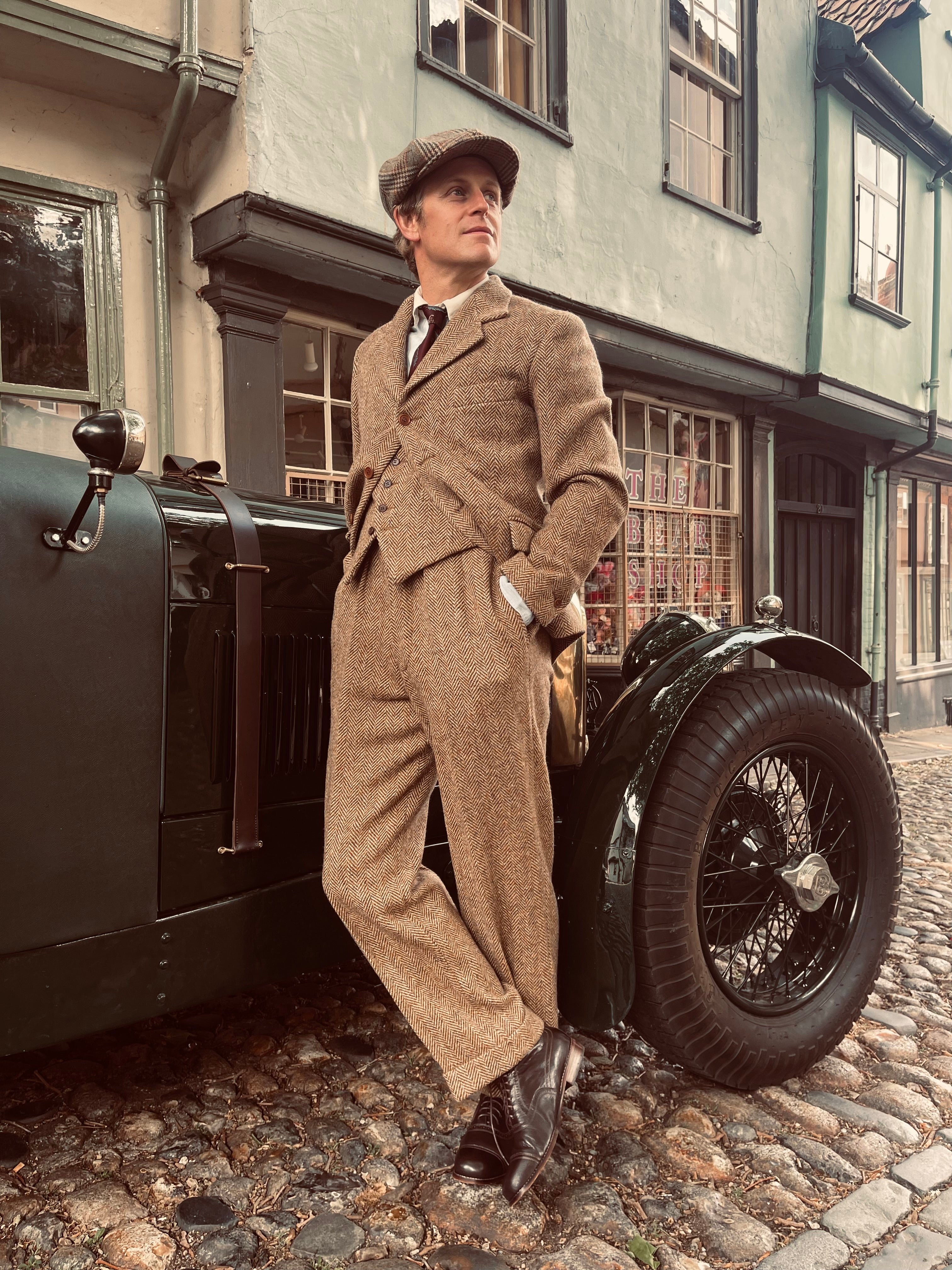 Man in vintage attire standing next to an old-fashioned car on a cobblestone street.