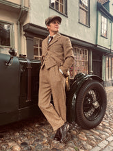 Man in vintage attire standing next to an old-fashioned car on a cobblestone street.