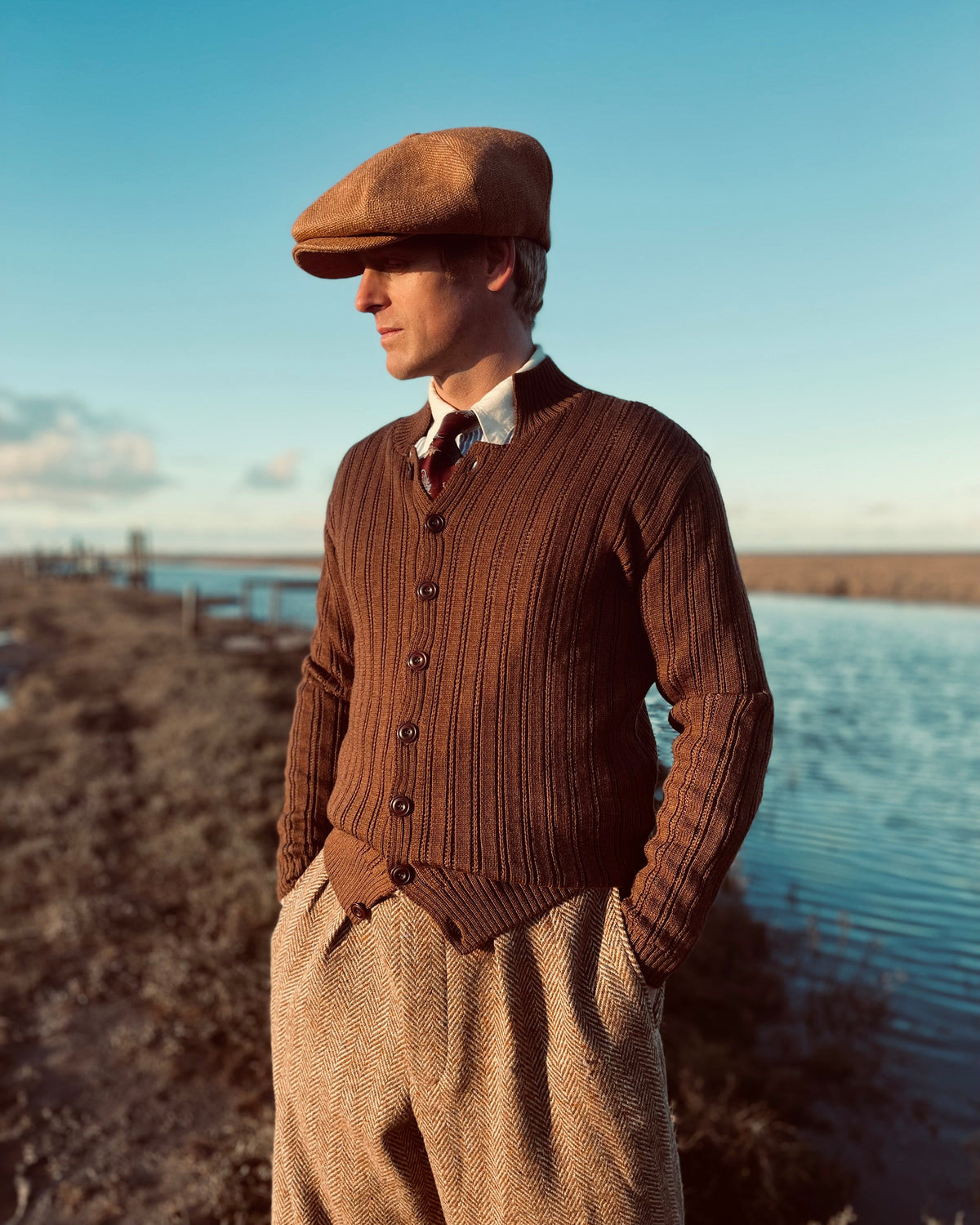 A man wearing a brown merino wool cardigan with raised ribbed stitch and Corozo buttons, paired with a shirt and tie, standing beside a body of water.