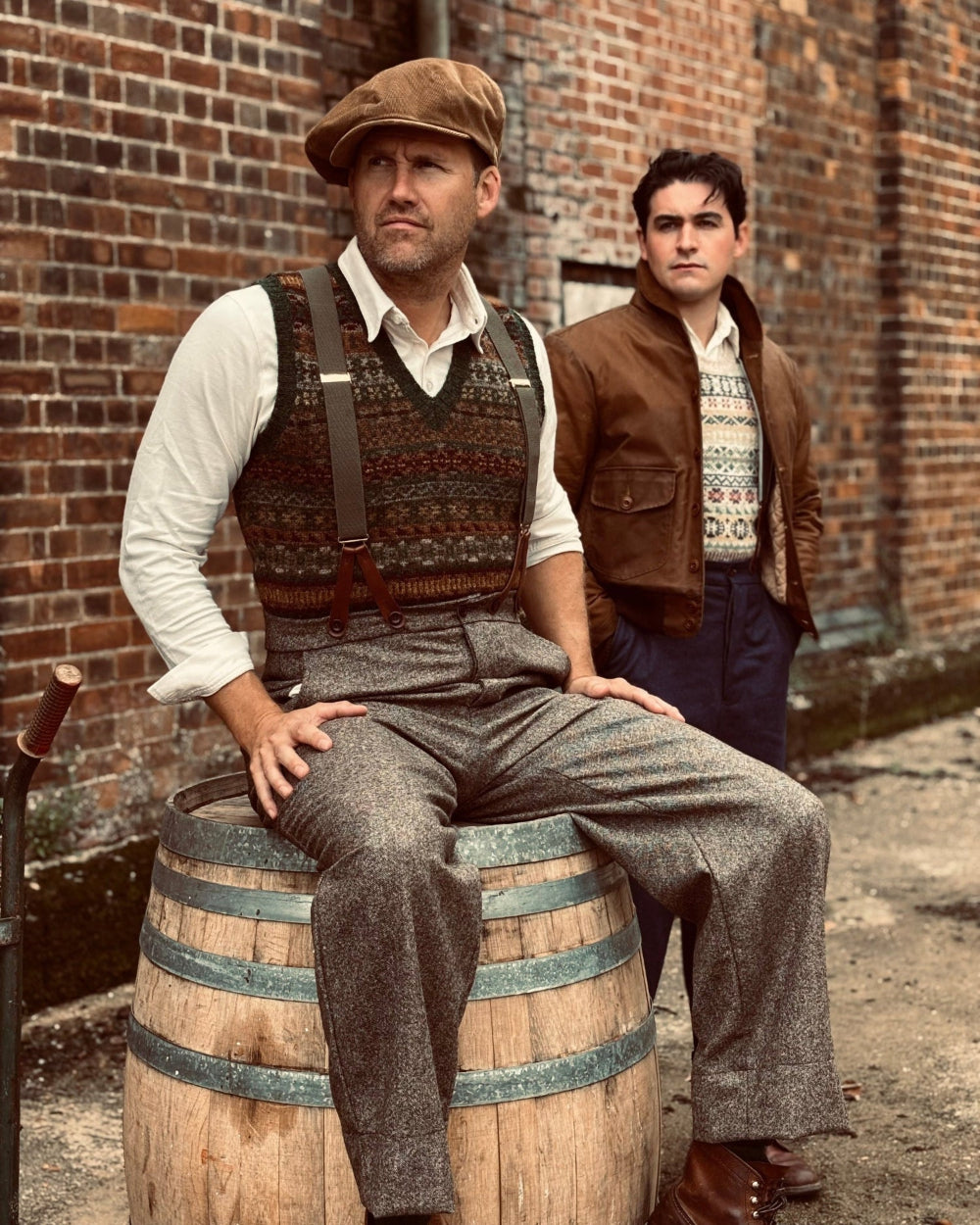 Two men dressed in vintage 1930s style clothing pose in front of a brick wall. The man in the foreground sits on a wooden barrel wearing a brown flat cap, patterned sweater vest, white shirt, suspenders, and gray tweed trousers.