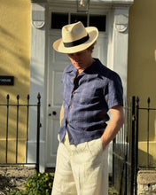 Man wearing a Panama hat and blue 1930s shirt standing in front of a white door.
