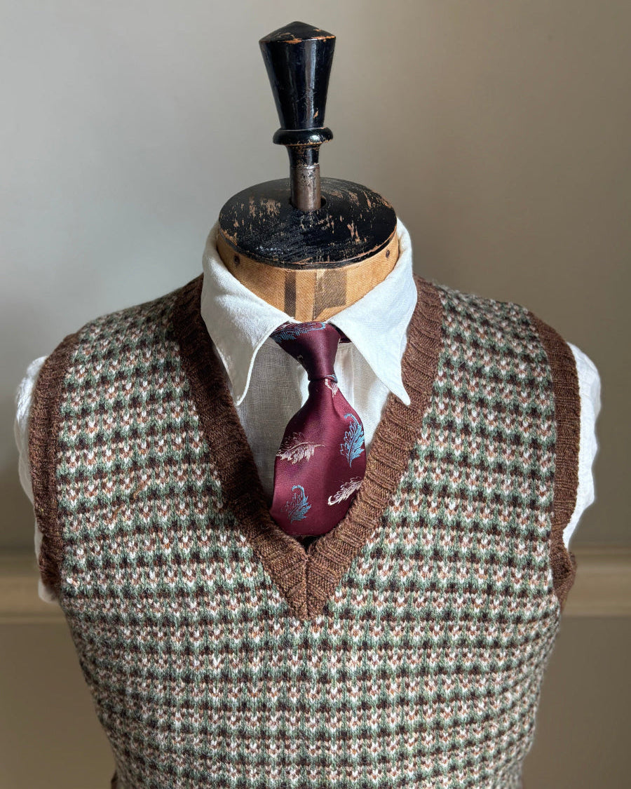 A brown and beige houndstooth knit sweater vest displayed on a wooden dress form mannequin, styled with a white collared shirt and a burgundy patterned necktie against a neutral beige background.