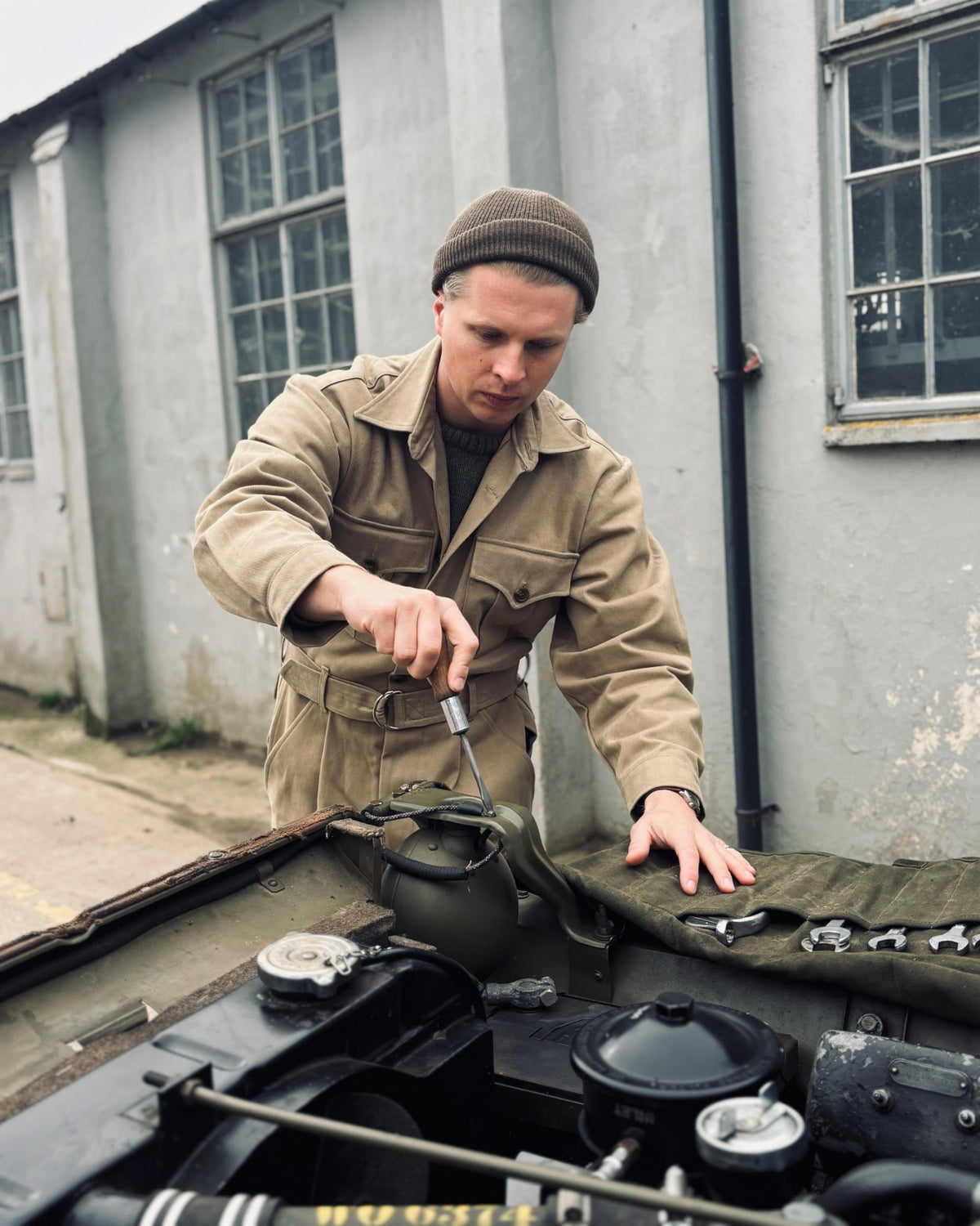 A man in a tan military-style jacket and brown beanie works on a vehicle engine outdoors in front of a gray building.