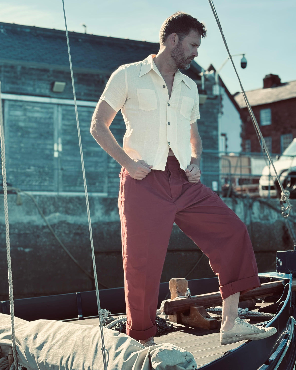 A man stands on the deck of a boat in a harbor, wearing a linen 1930s cream short-sleeve button-up shirt and dusty rose cropped trousers.