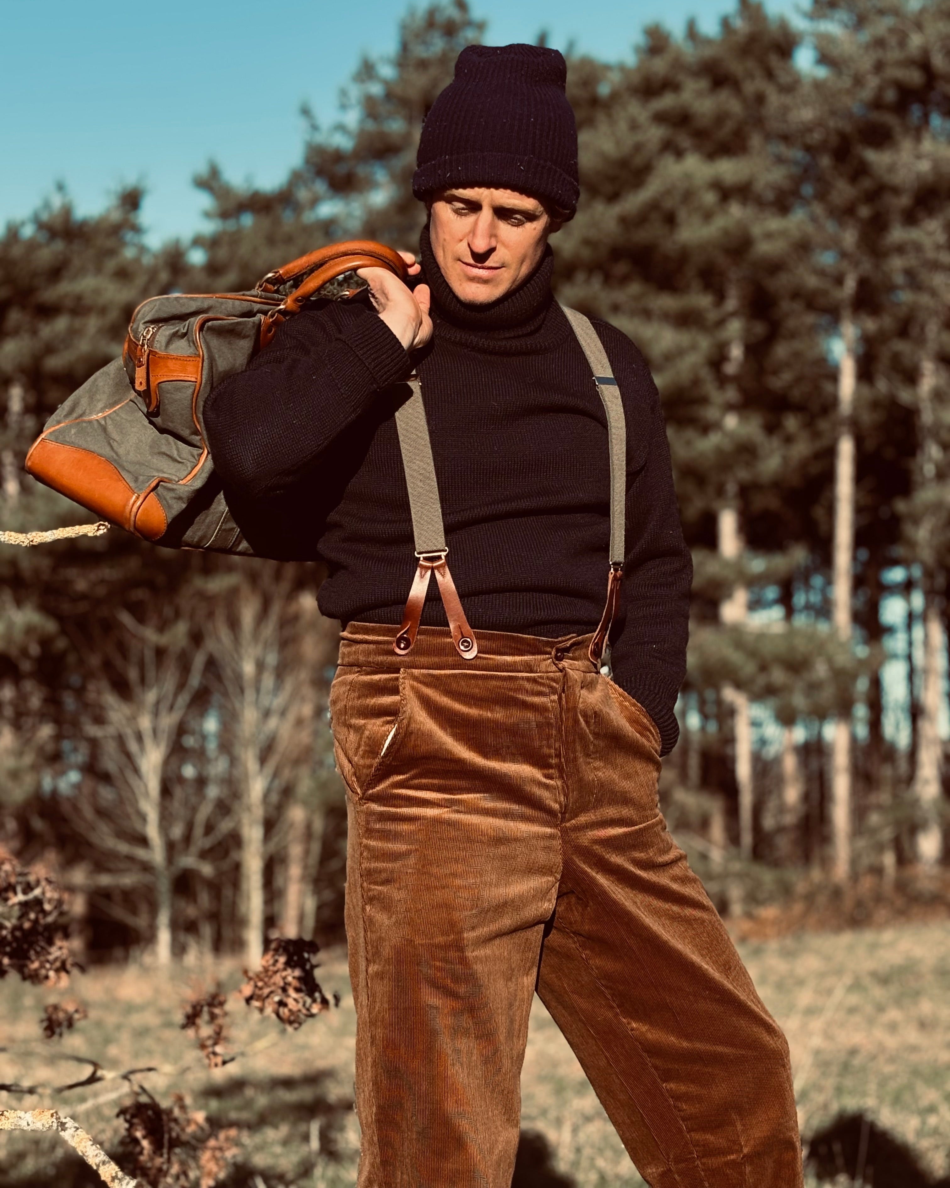 Man in brown overalls and black beanie holding a backpack in a forest setting