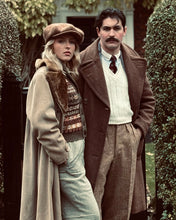 Two people in vintage clothing standing outdoors with greenery in the background