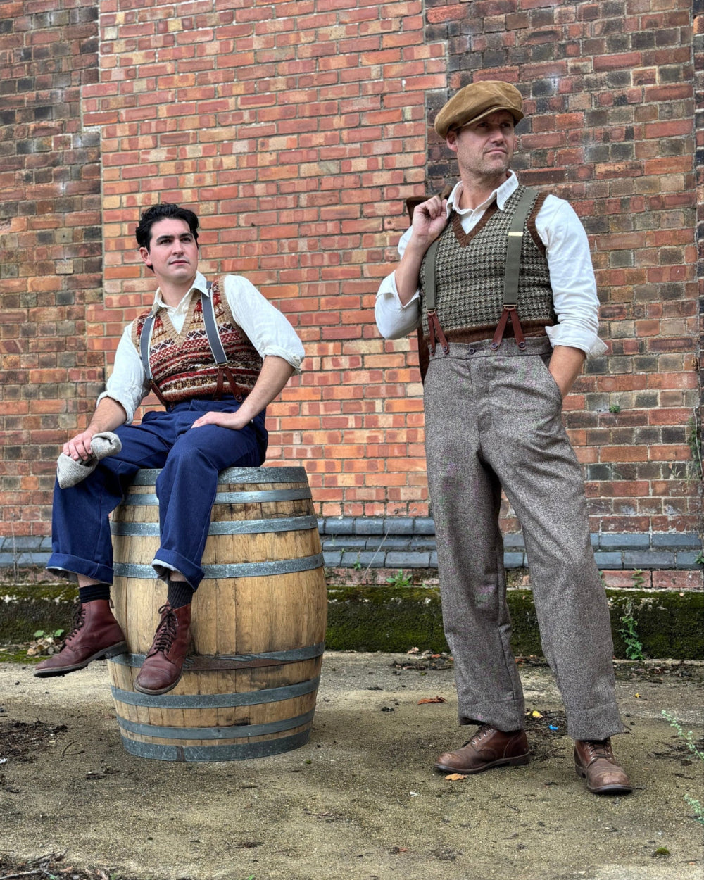 Two men in vintage-style clothing pose outdoors against a red brick wall. One man sits on a wooden barrel wearing a white shirt, patterned vest, blue trousers, and brown boots, while the other stands wearing a brown flat cap, tweed vest, white shirt, gray herringbone trousers, and brown boots.  Wide legged vintage trousers.