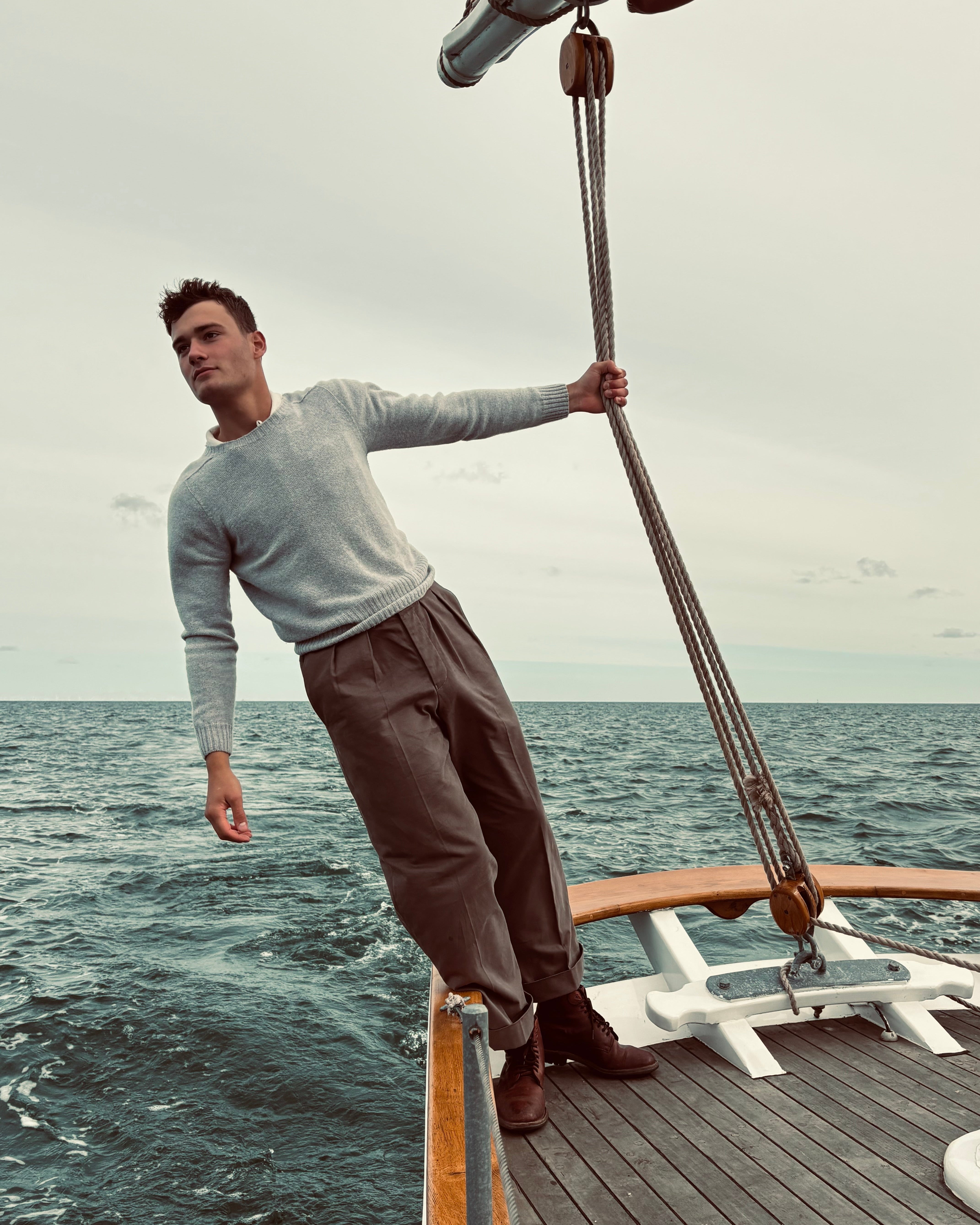 A man stands on the deck of a boat holding a rope, wearing a light gray long-sleeve knit sweater and dark brown trousers, with the ocean and overcast sky in the background.