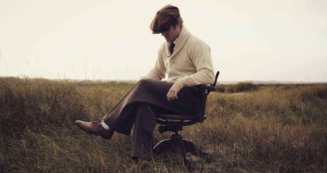 A man wearing a brown flat cap, cream sweater, brown trousers, and brown shoes sits on a black office chair in an open grassy field.