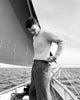 Man on a boat wearing a light sweater and dark pants, with water and sailboat in the background.