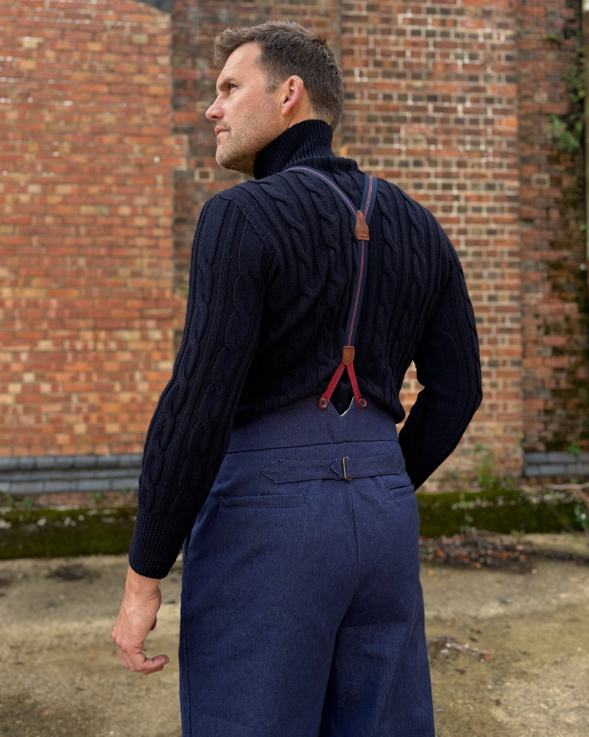 A man wearing a dark navy cable-knit turtleneck sweater and matching navy trousers with red suspenders, photographed outdoors against a red brick wall.